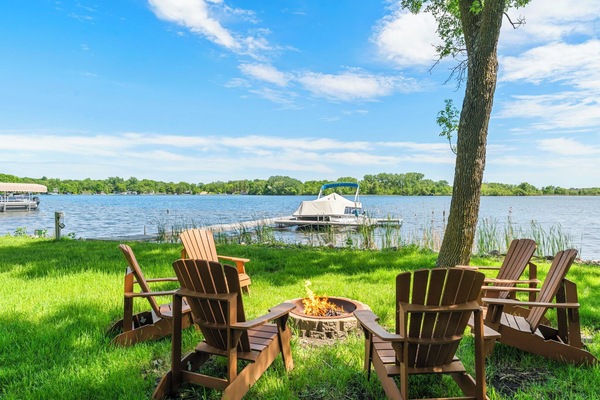 Private lakefront setting with Adirondack chairs around a fire pit. The property includes a boat dock on calm waters, surrounded by lush trees and natural shoreline.