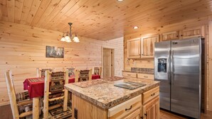 Interior de una cabaña de madera con cocina, mesa de comedor de madera y sillas, y un refrigerador.