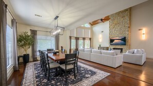 Uma sala de estar ampla com lareira de pedra, área de jantar com mesa e cadeiras de madeira, e um sofá branco grande.