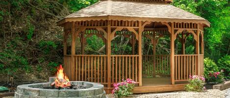 A wooden gazebo with a fire pit in front, surrounded by greenery.