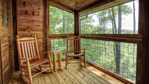 Una cabaña de madera con dos mecedoras y una pequeña mesa, ventanas amplias con vistas a un bosque.