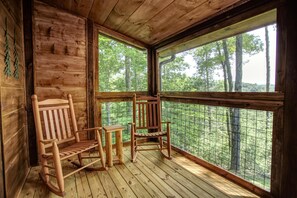 Eine Holzdiele mit zwei Schaukelstühlen und einem kleinen Tisch, große Fenster mit Blick auf einen Wald.