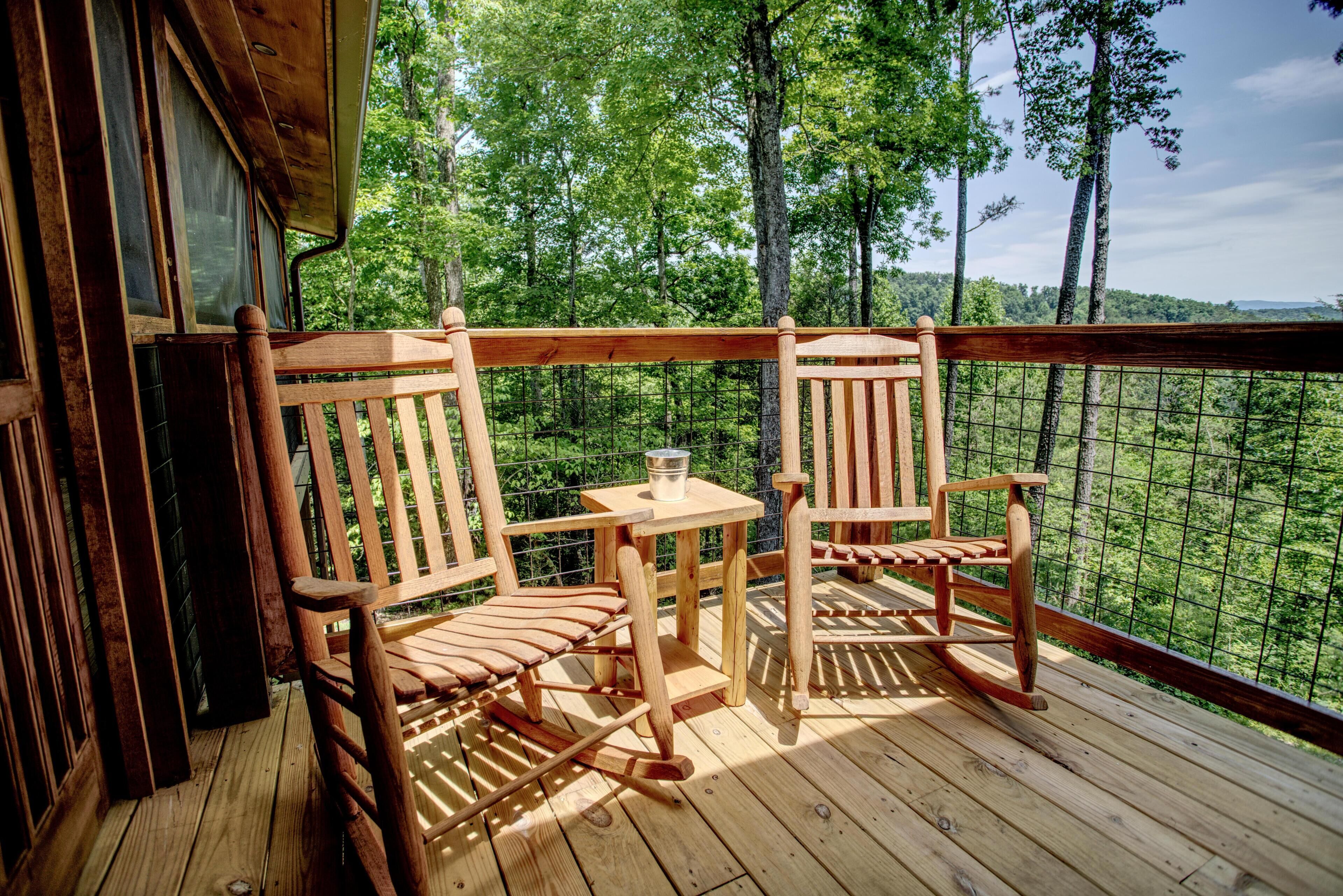 Een houten terras met twee schommelstoelen en een kleine tafel, met uitzicht op een bebost gebied.