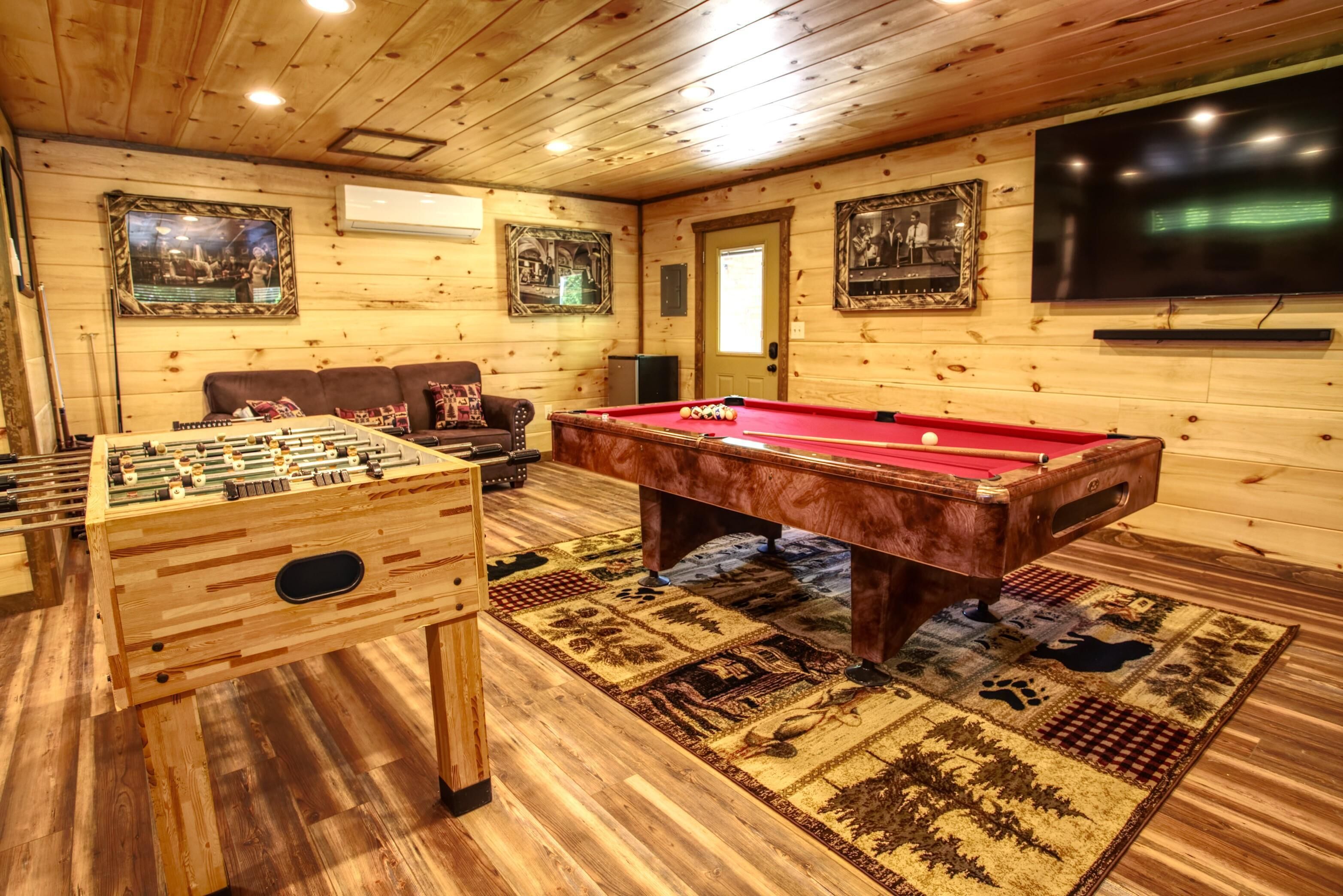Une pièce au plancher en bois, équipée d’une table de billard, d’une table de baby-foot et ornée de deux tableaux encadrés au mur.