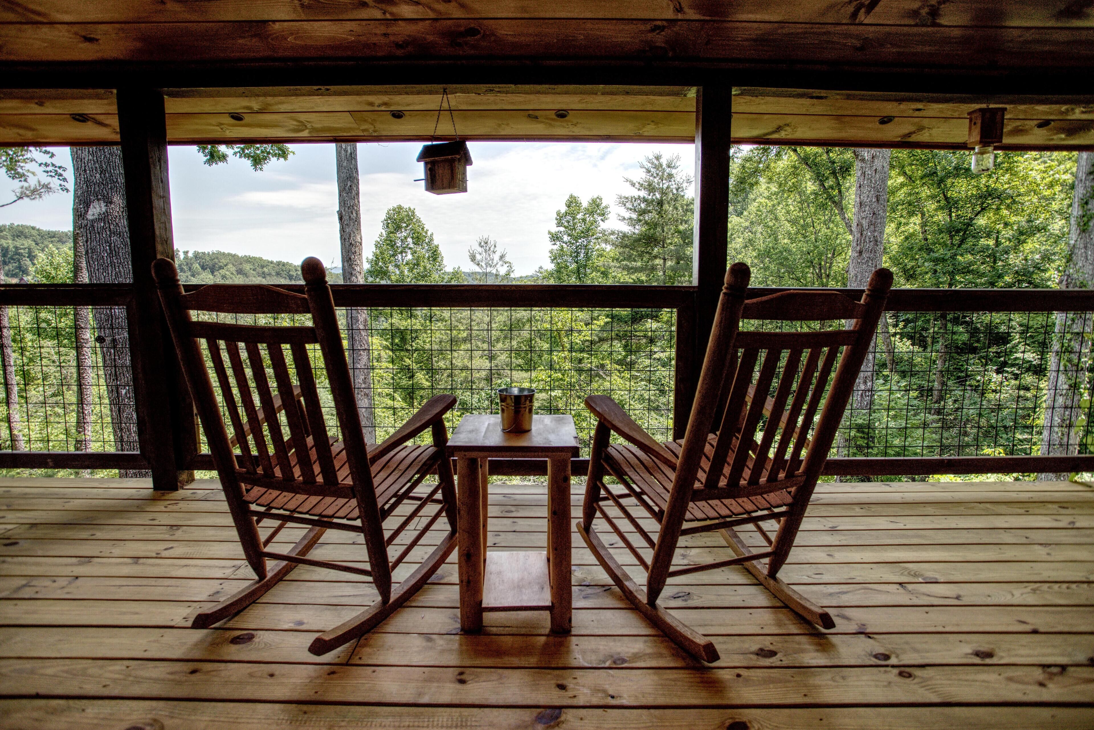 A wooden deck with two rocking chairs and a table, overlooking a forested area.