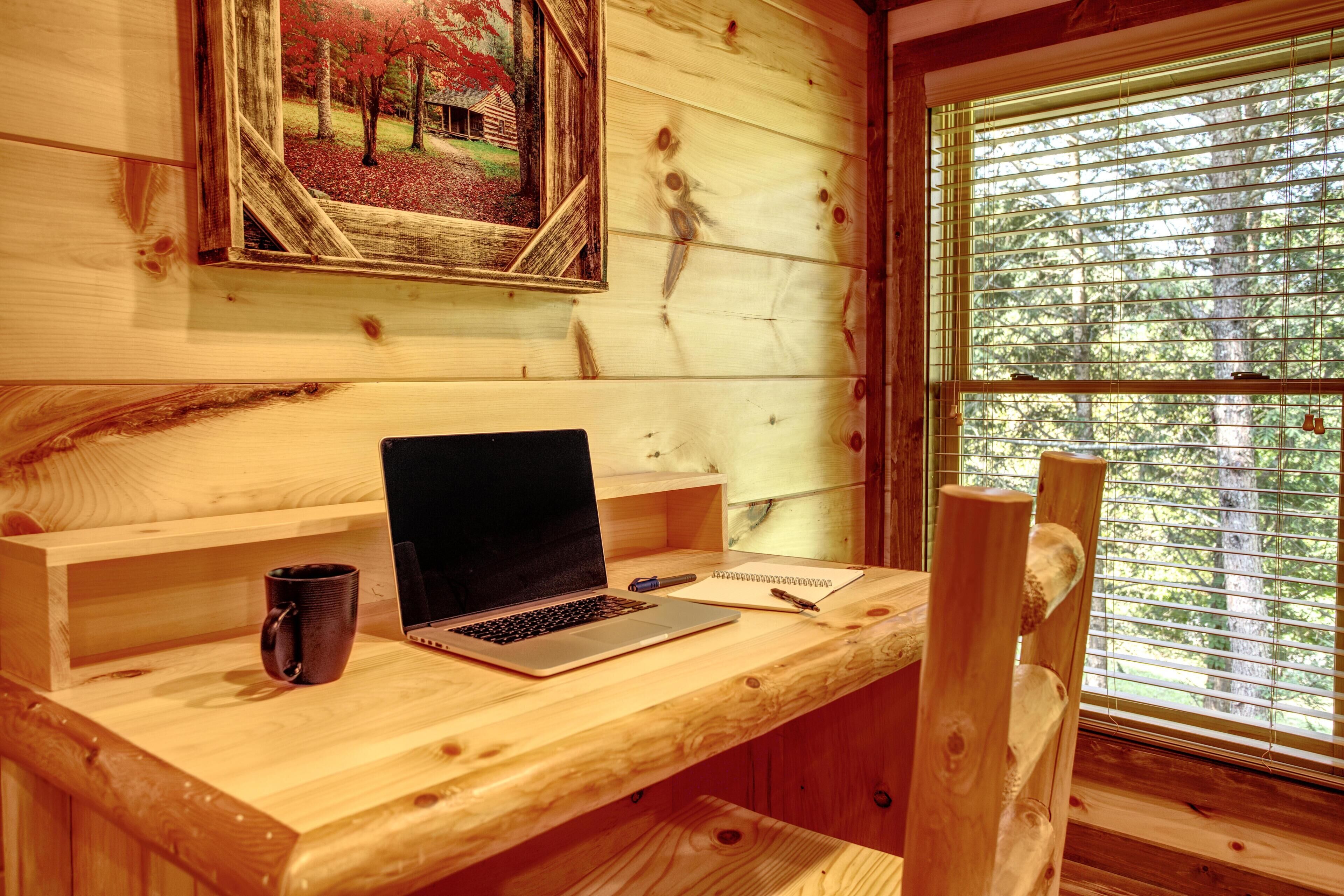 Un escritorio de madera con una computadora portátil, un cuaderno y un bolígrafo, un cuadro en la pared y una ventana con persianas.
