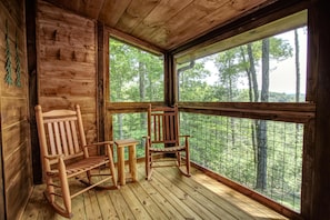 Eine Holzdiele mit zwei Schaukelstühlen und einem kleinen Tisch, große Fenster mit Blick auf einen Wald.