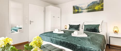 Un dormitorio con cama, mesita de noche, lámpara, silla y un cuadro en la pared.