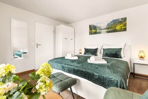 Un dormitorio con cama, mesita de noche, lámpara, silla y un cuadro en la pared.