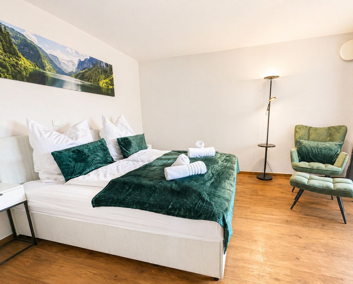 Ein ordentlich bezogenes Bett mit grüner Decke, ein grüner Sessel, eine Stehlampe und ein Gemälde einer Berglandschaft an der Wand.