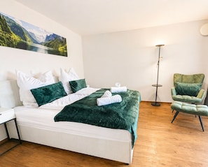 A neatly made bed with a green blanket, a green chair, a floor lamp, and a painting of a mountain landscape on the wall. - Suite 2 on the ground floor with balcony (Bad Aibling)