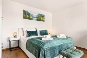 Un dormitorio con cama, almohadas, mesita de noche, lámpara, banco y un cuadro en la pared.