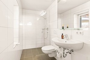 Un baño moderno con azulejos blancos, un inodoro, un lavabo, una ducha y un espejo.