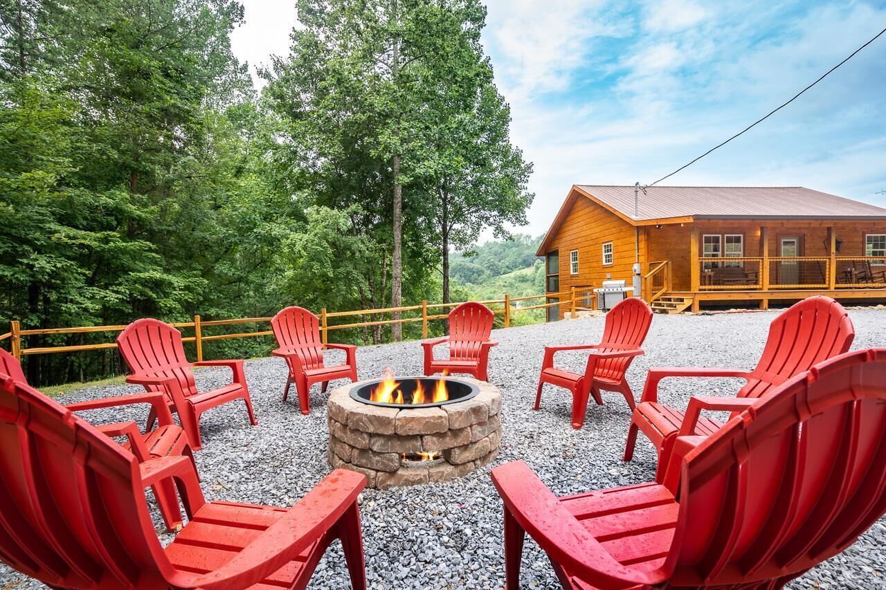 A fire pit surrounded by red Adirondack chairs, a wooden cabin, and a forested backdrop.