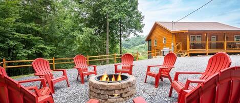 A fire pit surrounded by red Adirondack chairs, a wooden cabin, and a forested backdrop.