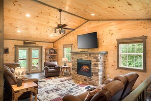 A log cabin interior with a fireplace, leather furniture, a television, and a view of the outdoors. - Tennessee Tranquility (Sevierville)