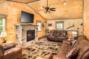 A log cabin interior with a stone fireplace, leather furniture, and a mounted television. - Tennessee Tranquility (Sevierville)