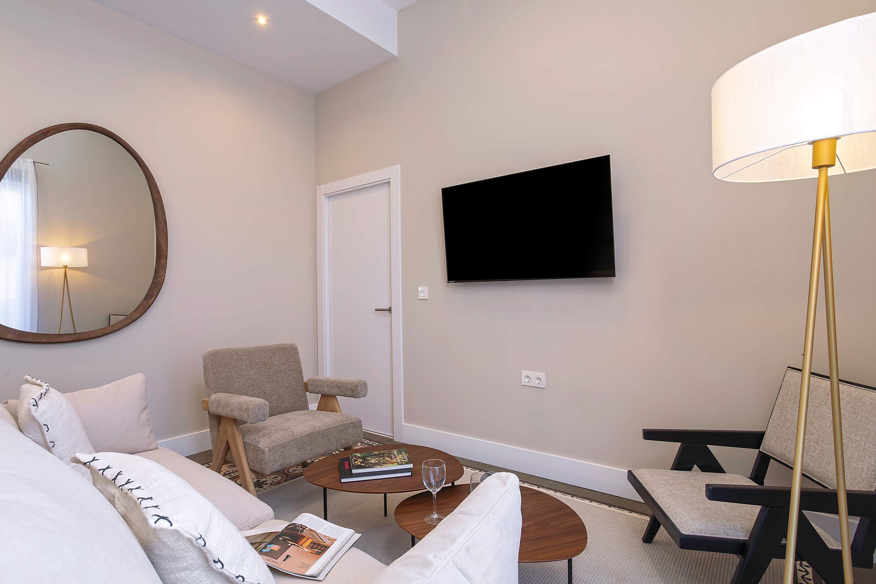 A modern living room with a sofa, two armchairs, a coffee table, a flat-screen TV, a round mirror, and a floor lamp.