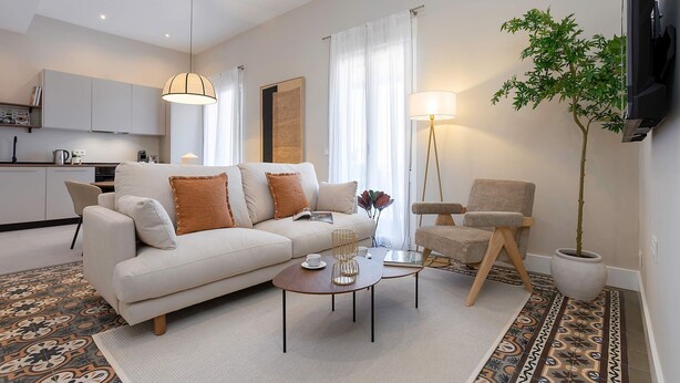 Ein modernes Wohnzimmer mit einer Couch, einem Couchtisch und einem Sessel, ein Essbereich mit Schränken und einer großen Topfpflanze.