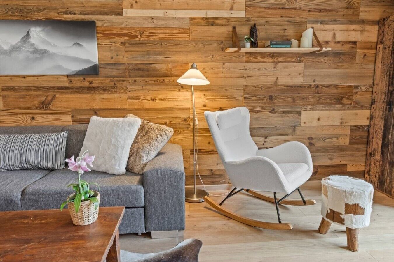 Ein gemütliches Zimmer mit einem grauen Sofa, einem weißen Schaukelstuhl, Holzboden und einem an der Wand befestigten Regal mit Büchern und Dekoration.