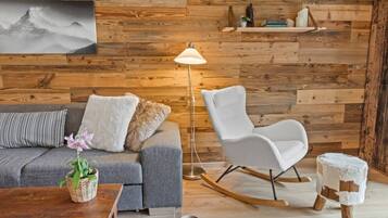 Ein gemütliches Zimmer mit einem grauen Sofa, einem weißen Schaukelstuhl, Holzboden und einem an der Wand befestigten Regal mit Büchern und Dekoration.