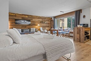 A bedroom with a large bed, a wooden wall, a dining area with a table and chairs, and a television on a wooden cabinet.
