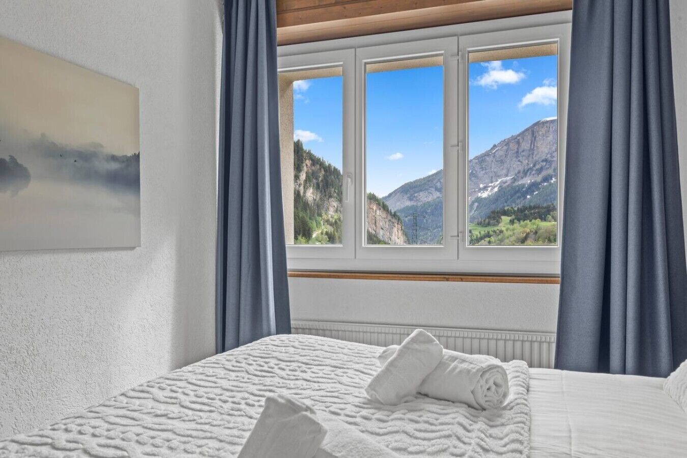 Quarto de hotel com cama, janela com vista para as montanhas e um quadro na parede.