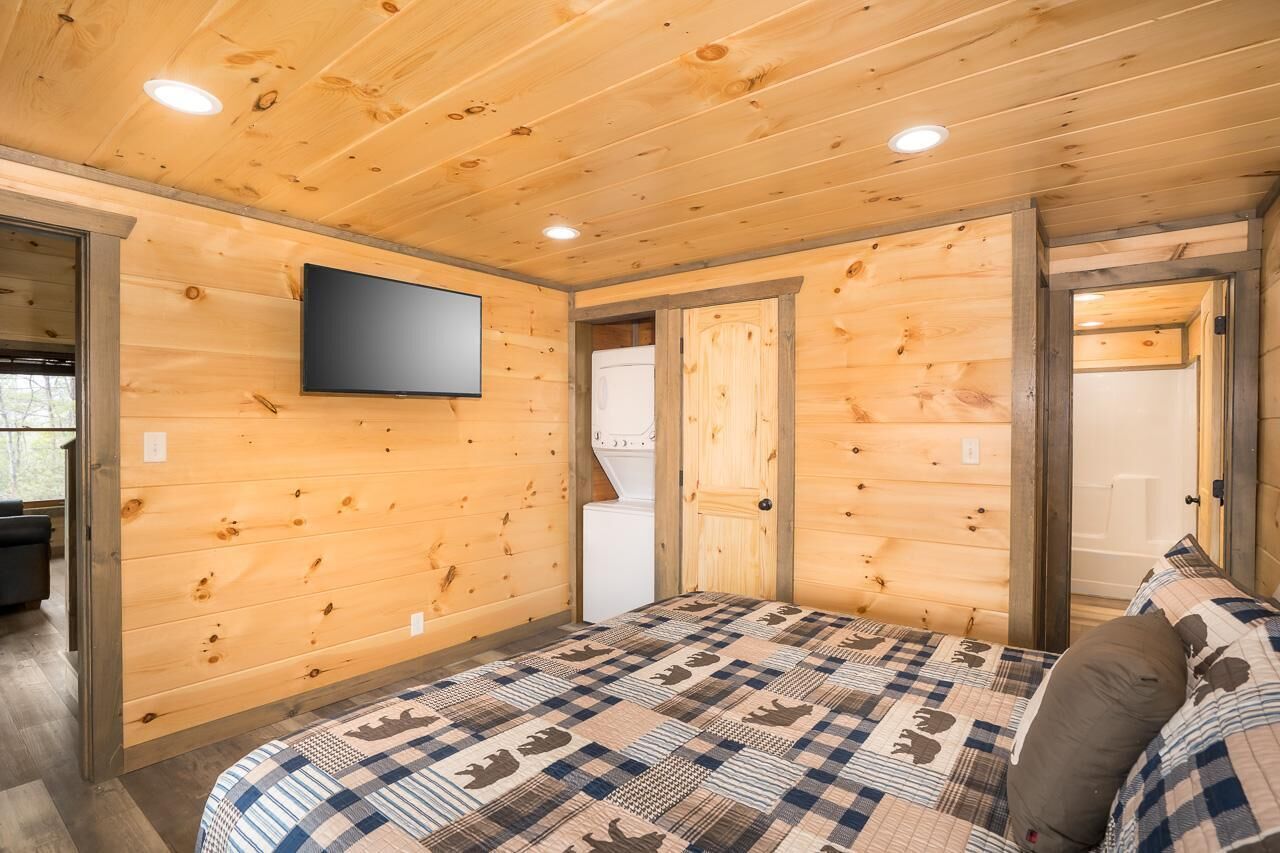 Habitación con paneles de madera, una cama, un televisor de pantalla plana y una puerta que da a otra habitación.