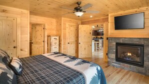 Ein Interieur einer Blockhütte mit einem Bett, einem Kamin, einem Deckenventilator und einem an der Wand montierten Fernseher.