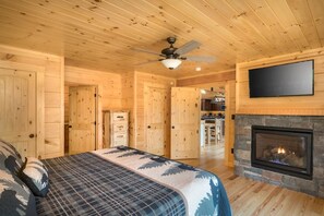 A log cabin interior with a bed, a fireplace, a ceiling fan, and a television mounted on the wall. - Four Bears (Gatlinburg)