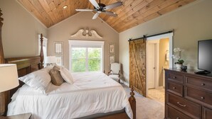 Een slaapkamer met een houten plafond, een bed met witte beddengoed, een nachtkastje, een dressoir, een televisie en een schuifdeur naar een andere kamer.