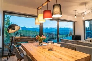 Een moderne eethoek met een houten tafel, stoelen en hanglampen. Grote ramen bieden uitzicht op bergen.