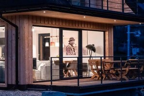 Een houten gevel van een gebouw met een glazen deur, een eetgedeelte met tafels en stoelen, en een zichtbaar interieur met een bank en lamp.
