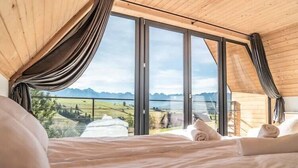 A bedroom with a large bed, wooden ceiling, and a view of a mountainous landscape through large windows.