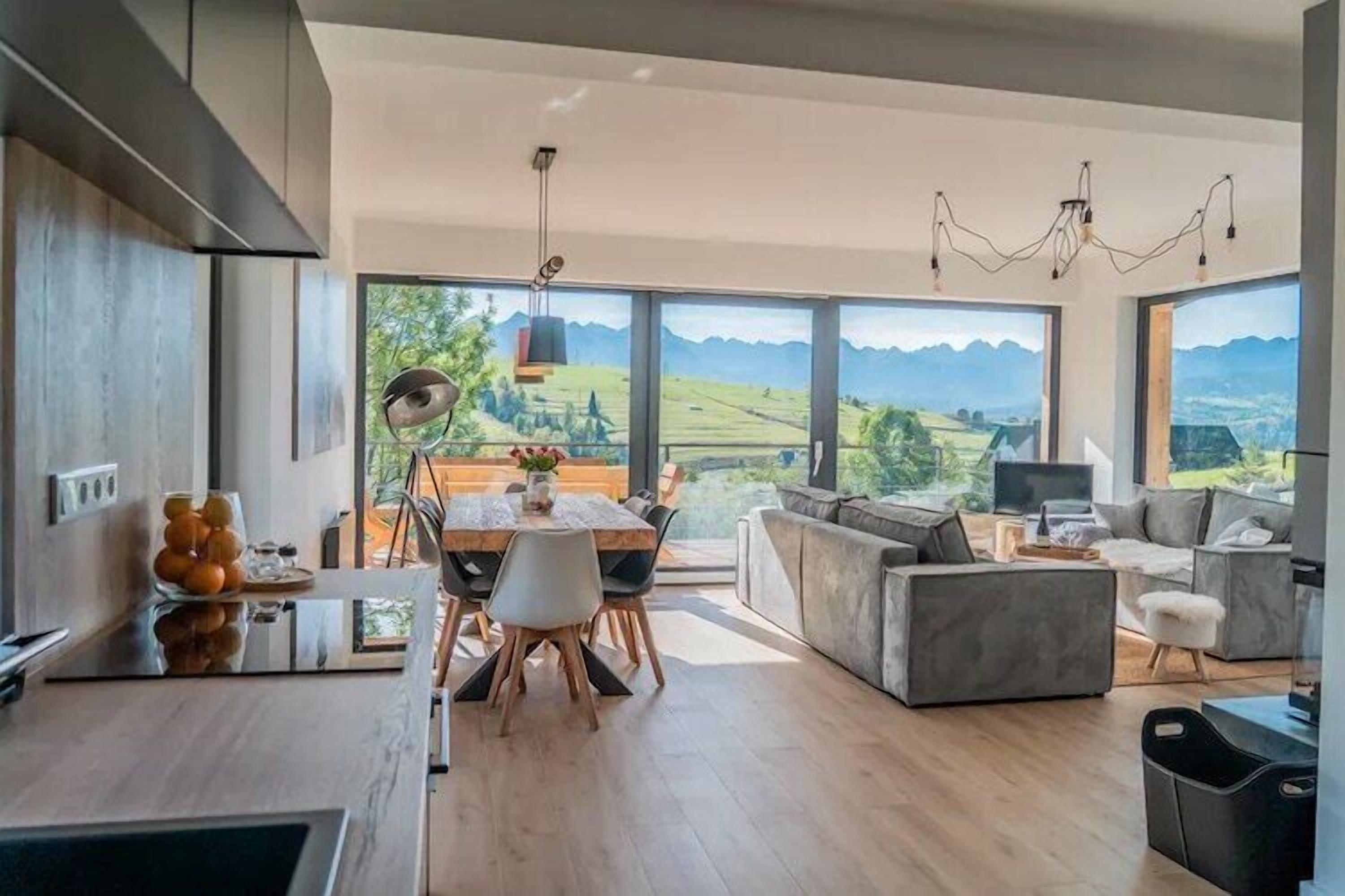 Une cuisine moderne avec un coin repas, de grandes fenêtres offrant une vue sur les montagnes, et un salon avec un canapé et un téléviseur.