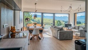 A modern kitchen with a dining area, large windows with a mountain view, and a living area with a sofa and a TV.