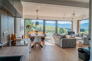 Una cocina moderna con comedor, ventanas amplias con vistas a la montaña y zona de estar con sofá y televisor.