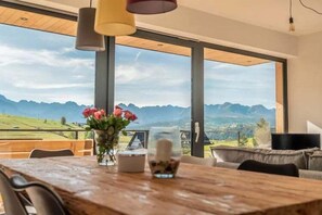 A dining area with a wooden table, chairs, a vase of flowers, and a large window offering a mountain view.