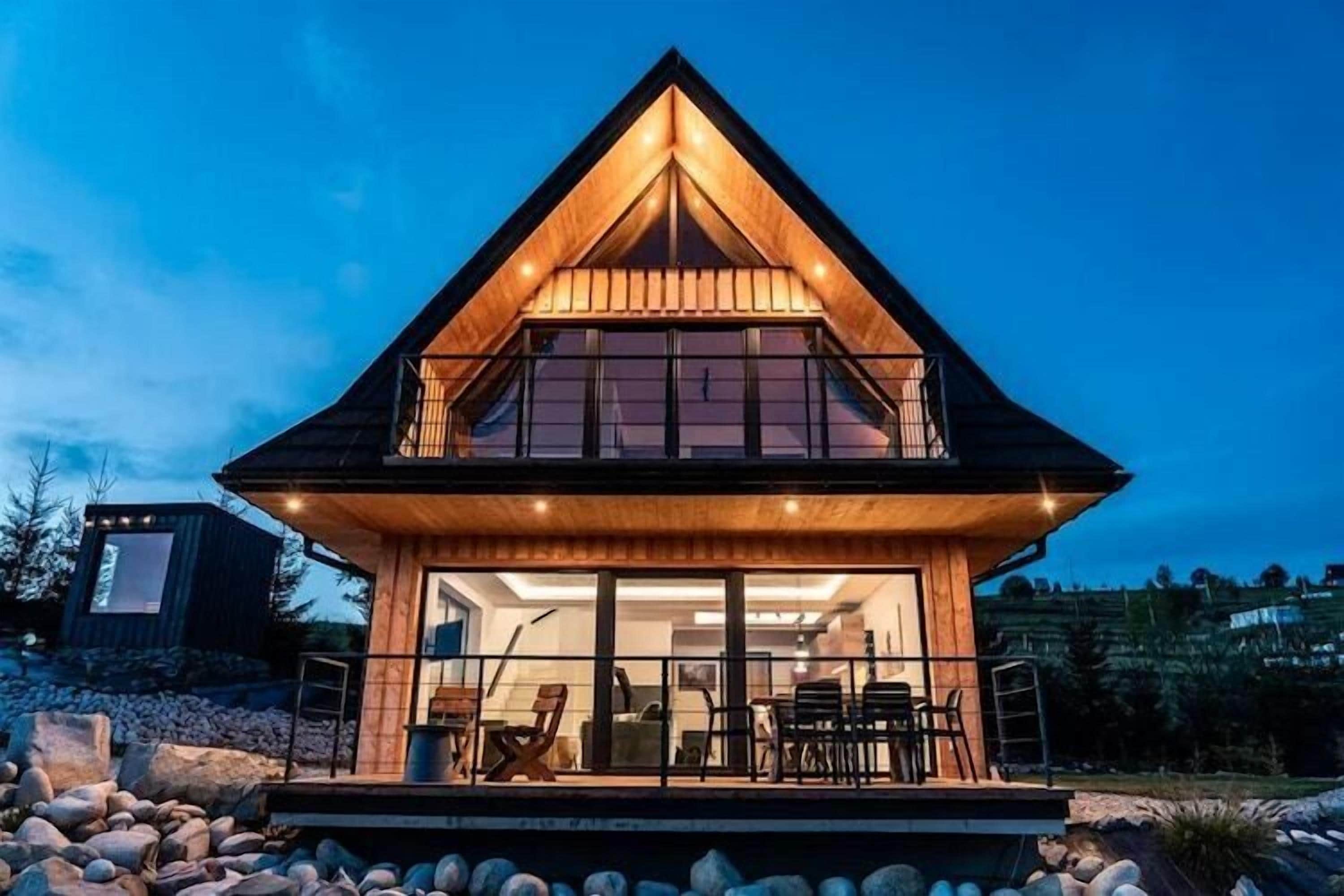 Une maison en bois à deux étages, dotée d’un balcon, de grandes fenêtres et d’un intérieur bien éclairé.
