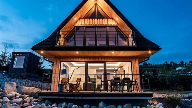 A two-story wooden house with a balcony, large windows, and a well-lit interior.