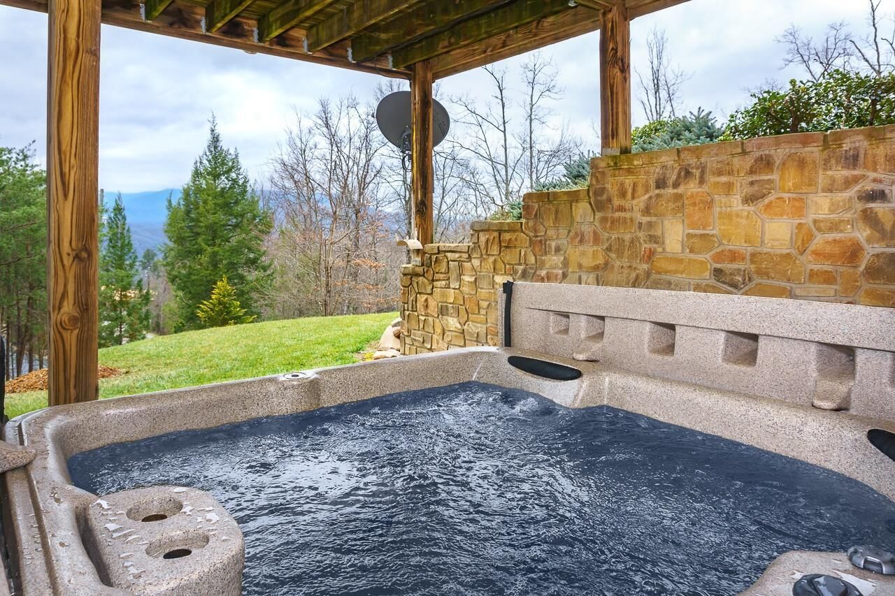 Uma banheira de hidromassagem com parede de pedra e pérgola de madeira, cercada por gramado e árvores.