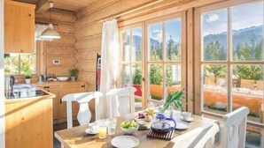 Una baita in legno con zona pranzo, cucina e ampie finestre che si affacciano su una vista sulle montagne.