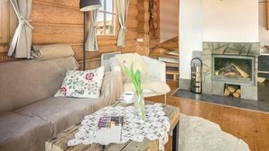 Un accogliente interno di una baita con un divano di legno, un tavolo con tovaglia di pizzo, un vaso con fiori e un camino.