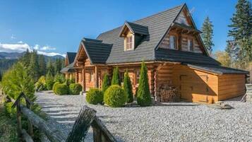 A log cabin with a gravel driveway, surrounded by greenery and mountains.