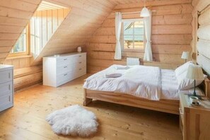 A cozy attic bedroom with a wooden sloped ceiling, a bed, a dresser, and a window with curtains.