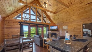A wooden cabin interior with a dining area, kitchen, fireplace, and large windows. - Another Day in Bearadise (Gatlinburg)
