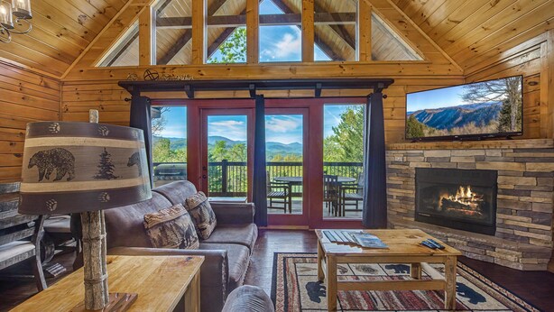 A log cabin interior with a fireplace, a sofa, a coffee table, and a view through the glass doors. - Another Day in Bearadise (Gatlinburg)
