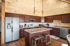A kitchen with wooden ceiling, stainless steel appliances, granite countertops, and a central island with stools. - Bear and Trout (Gatlinburg)