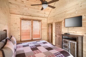 Quarto de cabana de madeira com uma cama, um ventilador de teto, uma TV e uma janela com persianas.