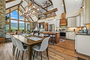 Un vaste espace de salle à manger avec une grande table et des chaises en bois, une cuisine dotée de meubles blancs et une vue sur les montagnes grâce à de grandes fenêtres.